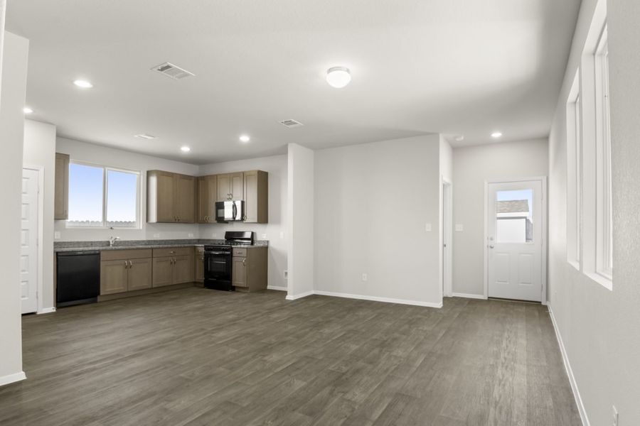 Image of a living room area with brown vinyl flooring and cream walls with a L-shaped kitchen in the corner