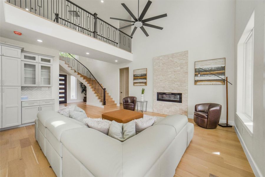 This spacious living room features a modern design with a high ceiling and a large ceiling fan. It includes a cozy fireplace, a stylish open staircase, and a comfortable seating area. Natural light floods the space through a large window, enhancing the warm, inviting atmosphere.
