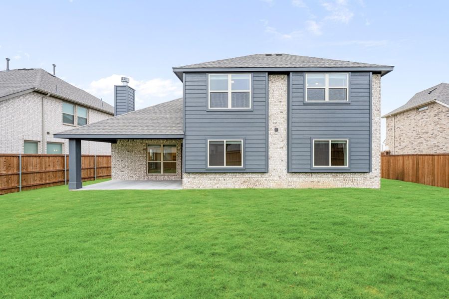 Two-story home rear view with gray siding, brick accents, covered patio, and fenced backyard with green lawn Two-story home rear view with gray siding, brick accents, covered patio, and fenced backyard with green lawn