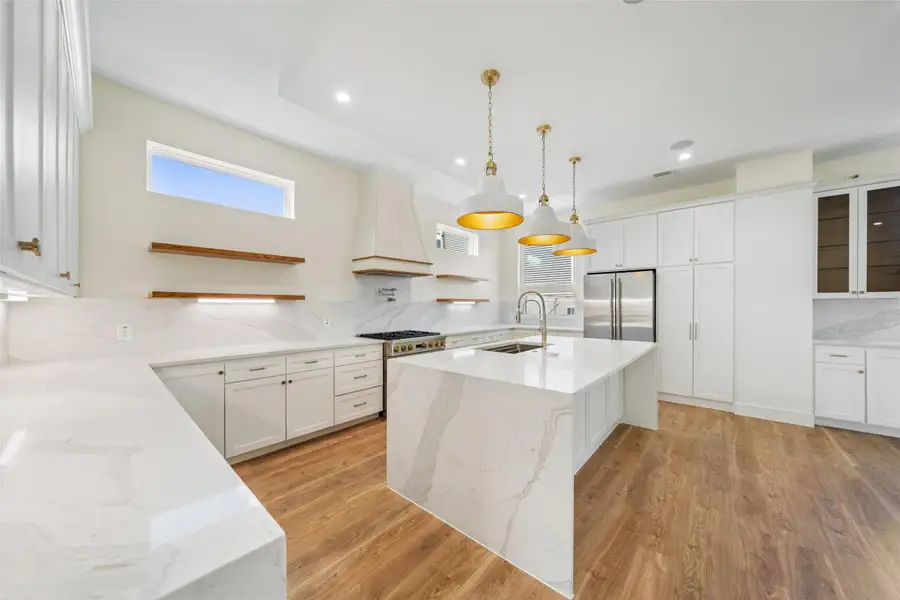 Kitchen features a modern design with white cabinetry, marble countertops, and a spacious island with a sink. It includes wood floors, pendant lighting, stainless steel appliances, and open shelving, offering a sleek and functional space.