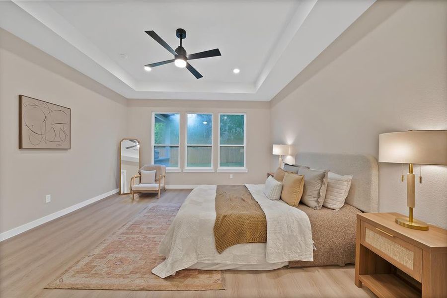 Spacious bedroom featuring light wood-finish flooring, a tray ceiling with a modern ceiling fan, recessed lighting, and three large windows Spacious bedroom featuring light wood-finish flooring, a tray ceiling with a modern ceiling fan, recessed lighting, and three large windows