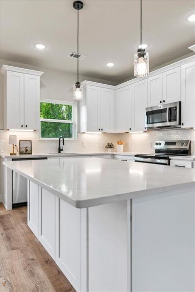 Bright kitchen featuring white shaker cabinetry, stainless steel appliances, light-toned countertops, wood-finish flooring, and pendant lighting