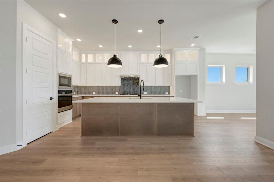 Kitchen with glass fronted cabinets, an island with sink, two tone color scheme, tasteful backsplash, and light wood finished floors