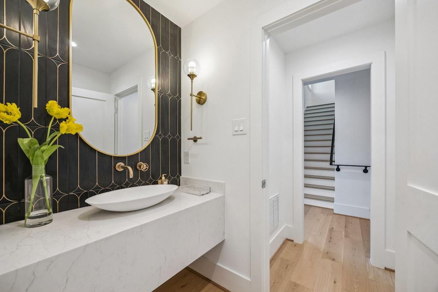 Second Floor Powder Room with a Floating Slab Quartz Counter, Oval Stone Composite White Vessel Sink, Wall-Mounted Widespread Faucet in Champange Bronze. The backsplash wall is adorned with 4 x 16 Art Deco Tile. There is also a Oval Framed Mirror and Two Bubble Glass Globe Wall Mounted Sconce Lights in Brass Gold.