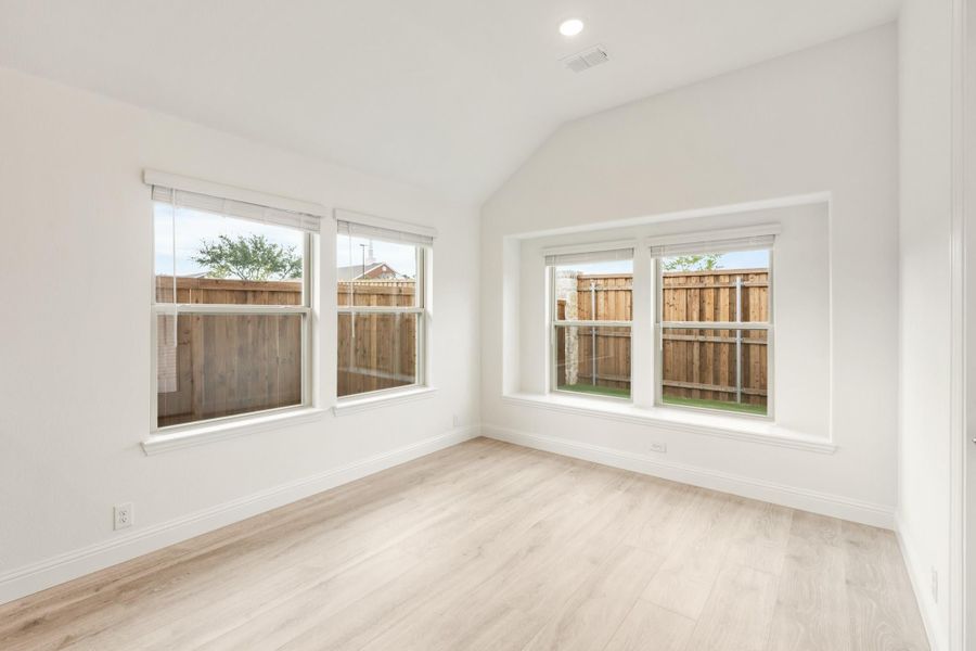 Empty bedroom with vaulted ceiling, light wood floors, and multiple windows overlooking a fenced backyard