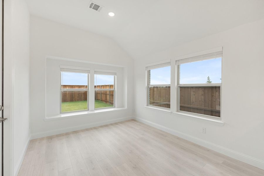 Empty bedroom with vaulted ceiling, light wood floors, and multiple windows overlooking a fenced backyard