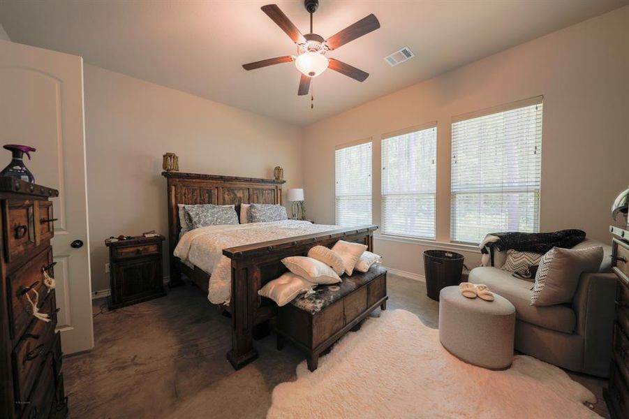 This cozy bedroom features a rustic wooden bed, soft lighting, and large windows that let in plenty of natural light. It includes a comfortable seating area, a ceiling fan, and warm, neutral tones. Ideal for relaxation.