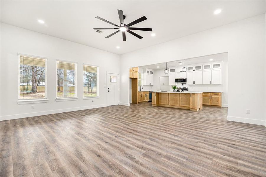 This photo showcases a spacious open-concept living area with modern wood flooring, a large ceiling fan, and abundant natural light from multiple windows. The kitchen features a central island, stylish pendant lighting, and a mix of white and wooden cabinetry, providing a sleek and inviting atmosphere.