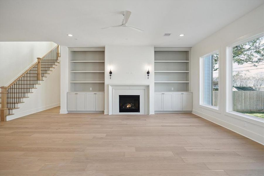 This stunning living room features a harmonious blend of style and functionality. Anchored by a sleek gas fireplace with a minimalist surround, the space exudes warmth and elegance. Flanking the fireplace are custom built-ins with open shelving and lower cabinets, offering both display and storage options.  A staircase with wood treads and iron balusters adds a touch of sophistication, seamlessly connecting this inviting space to the rest of the home.