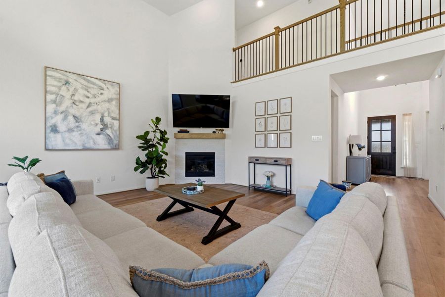 This inviting living room features high ceilings and an open layout with a cozy fireplace. Wood look vinyl plank flooring Loft-style railing above enhances the space. Natural light fills the room from the adjacent entryway.