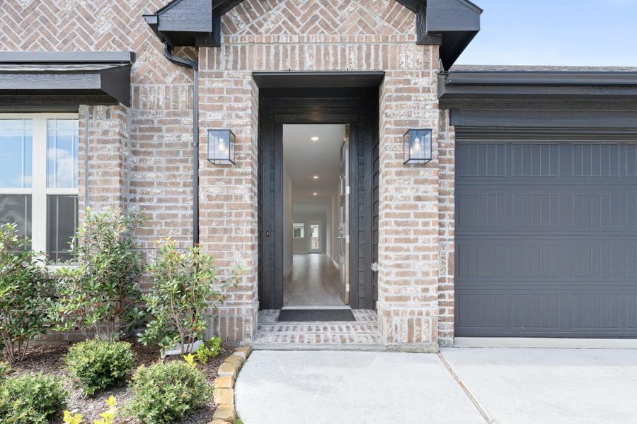Brick home entrance with open black front door, wall sconces, and gray garage door nearby
