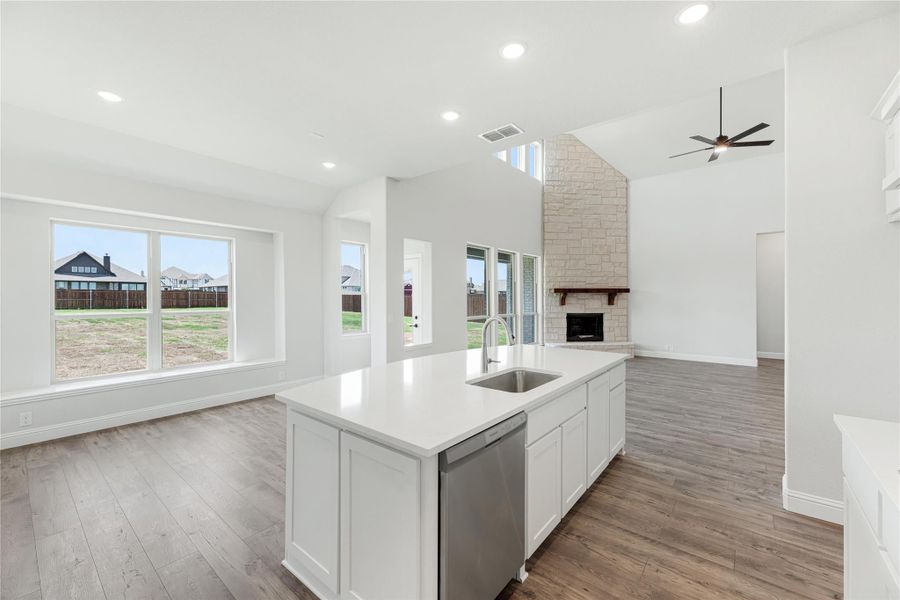 Open kitchen with white island, sink, and stainless dishwasher, opening to living area with stone fireplace and hardwood floors