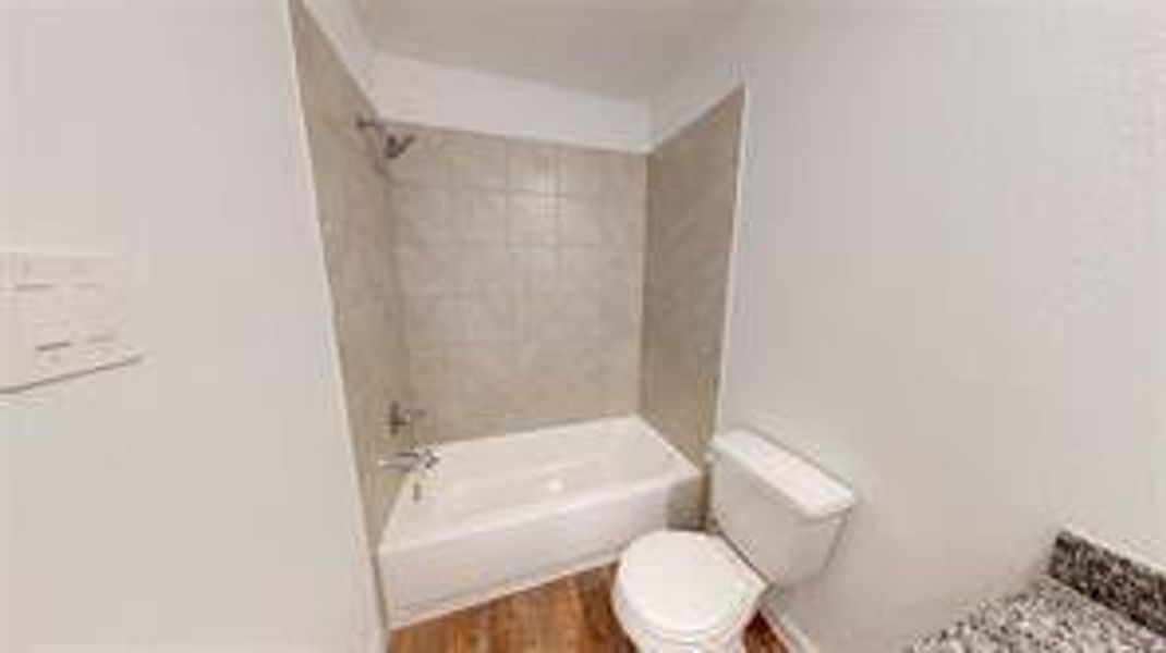 This image showcases a neatly designed bathroom within a floor plan, featuring a full-size bathtub with overhead shower, a toilet, and neutral-toned tiling. The wood like flooring adds warmth to the space, while the compact layout is efficiently structured for ease of use and functionality.