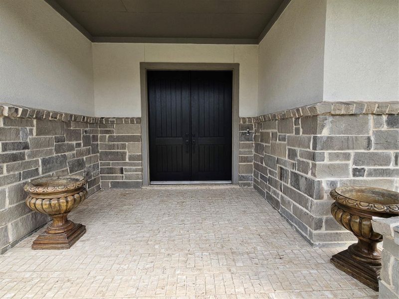 The photo showcases a welcoming front entrance with a stylish black double door framed by elegant stonework. The covered porch features decorative urns on either side, resting on a textured tile floor. The photo showcases a welcoming front entrance with a stylish black double door framed by elegant stonework. The covered porch features decorative urns on either side, resting on a textured tile floor.