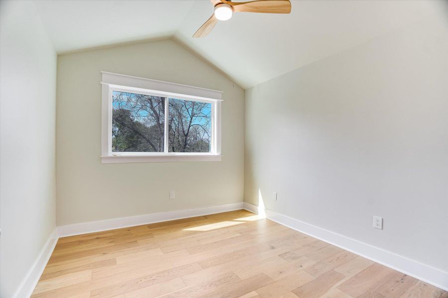 The room features light wood flooring, a window providing natural light and views of trees, and a vaulted ceiling with a ceiling fan The room features light wood flooring, a window providing natural light and views of trees, and a vaulted ceiling with a ceiling fan