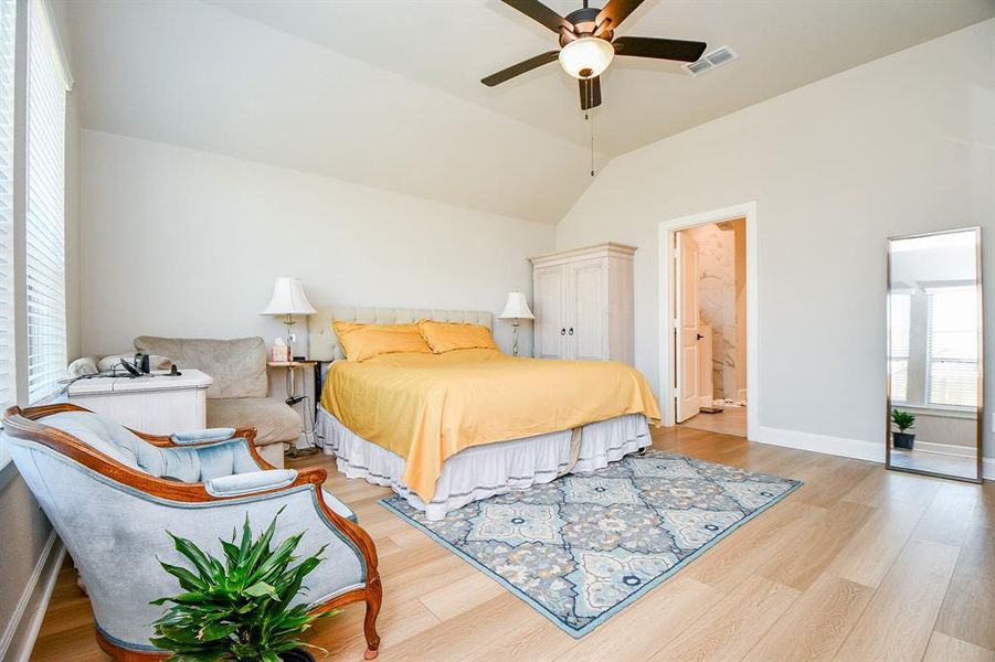 Bright and spacious bedroom with hardwood floors and vaulted ceiling. Features a cozy bed with yellow bedding, a stylish armchair, and a full-length mirror. Large windows provide ample natural light. Connected en-suite bathroom with modern fixtures.
