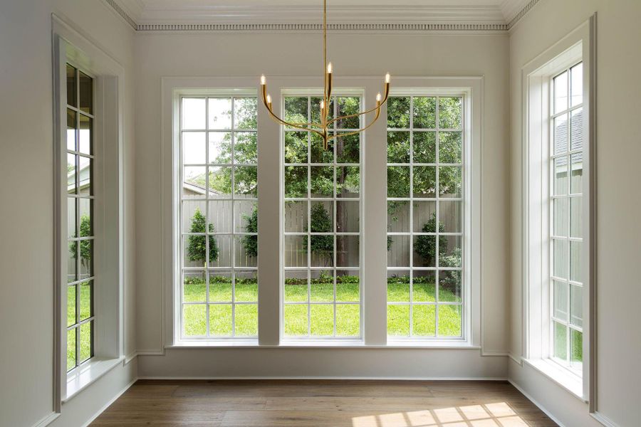 Sun-drenched breakfast room with a wall of oversized gridded Pella windows overlooking the landscaped backyard. Wide-plank hardwood floors, detailed dentil crown molding, a brass chandelier, and additional windows on both flanking walls flood the space with natural light. Sun-drenched breakfast room with a wall of oversized gridded Pella windows overlooking the landscaped backyard. Wide-plank hardwood floors, detailed dentil crown molding, a brass chandelier, and additional windows on both flanking walls flood the space with natural light.