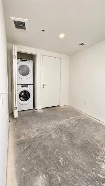 Stacked laundry unit with white appliances, enclosed within a closet featuring bi-fold doors, complemented by recessed ceiling lighting and a flat-panel interior door Stacked laundry unit with white appliances, enclosed within a closet featuring bi-fold doors, complemented by recessed ceiling lighting and a flat-panel interior door
