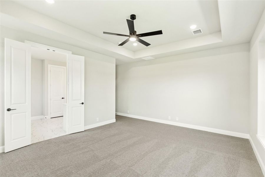 Empty bedroom with tray ceiling, black ceiling fan, gray carpet, and open doors to ensuite bathroom Empty bedroom with tray ceiling, black ceiling fan, gray carpet, and open doors to ensuite bathroom