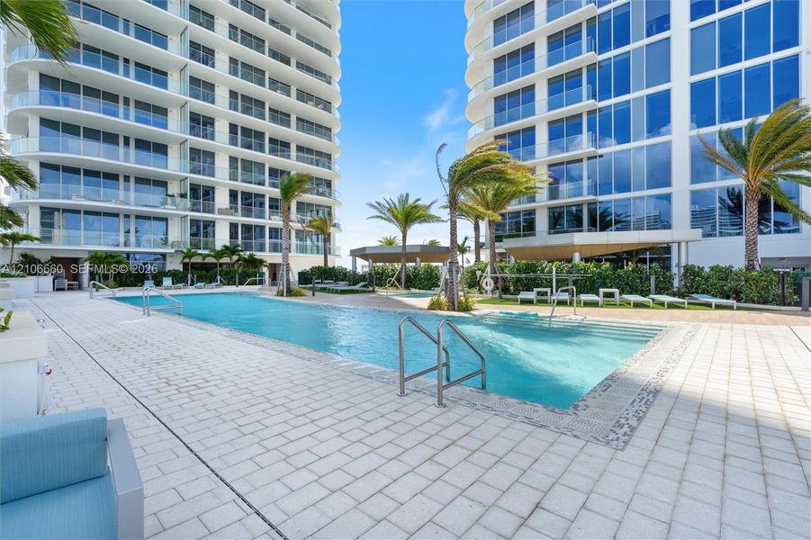Expansive outdoor swimming pool featuring stainless steel handrails, light-toned stone decking, mature palm trees, and structural shade pavilions