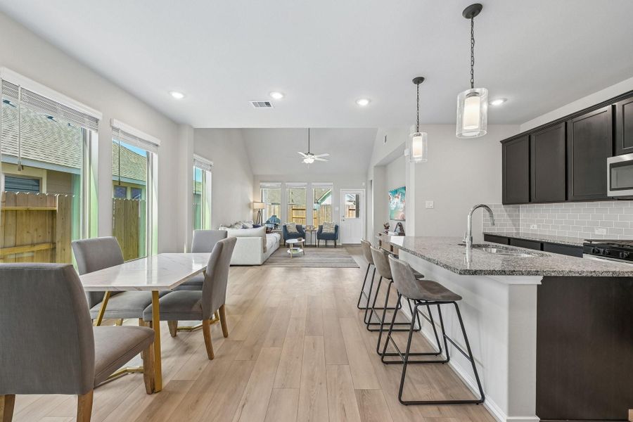 Bright and spacious open-concept living area with modern kitchen, featuring sleek dark cabinetry, granite countertops, and a large island with seating. The dining area and cozy living room are flooded with natural light from abundant windows, creating an inviting atmosphere.