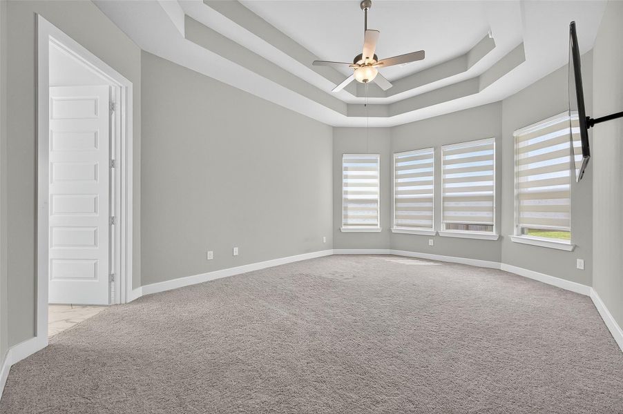 Luxurious primary suite showcases a dramatic double tray ceiling, bay window with modern shades, and plenty of space for a full bedroom set and sitting area.