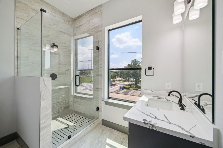 Bright and inviting, this bathroom showcases a walk-in shower with frameless glass and tile surround, paired with a vanity topped in quartz with clean-lined fixtures. A large window allows natural light to fill the space while offering a view toward the front of the property.