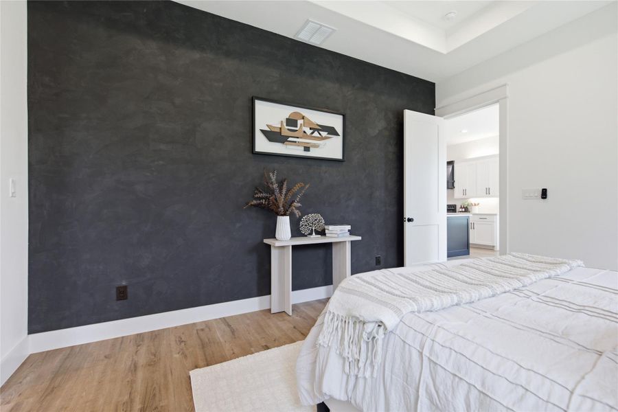 Well-lit sleeping quarters with a high tray ceiling and hardwood flooring, offering a glimpse of the adjoining kitchen. Well-lit sleeping quarters with a high tray ceiling and hardwood flooring, offering a glimpse of the adjoining kitchen.
