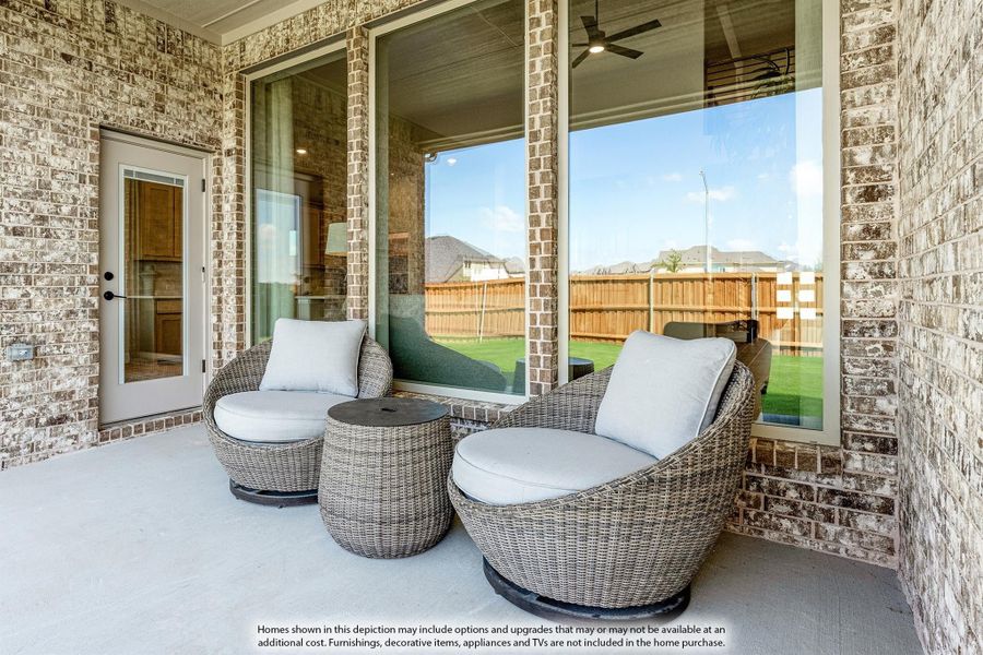 Covered brick patio with two wicker chairs, a small table, and large sliding glass doors overlooking a fenced backyard.