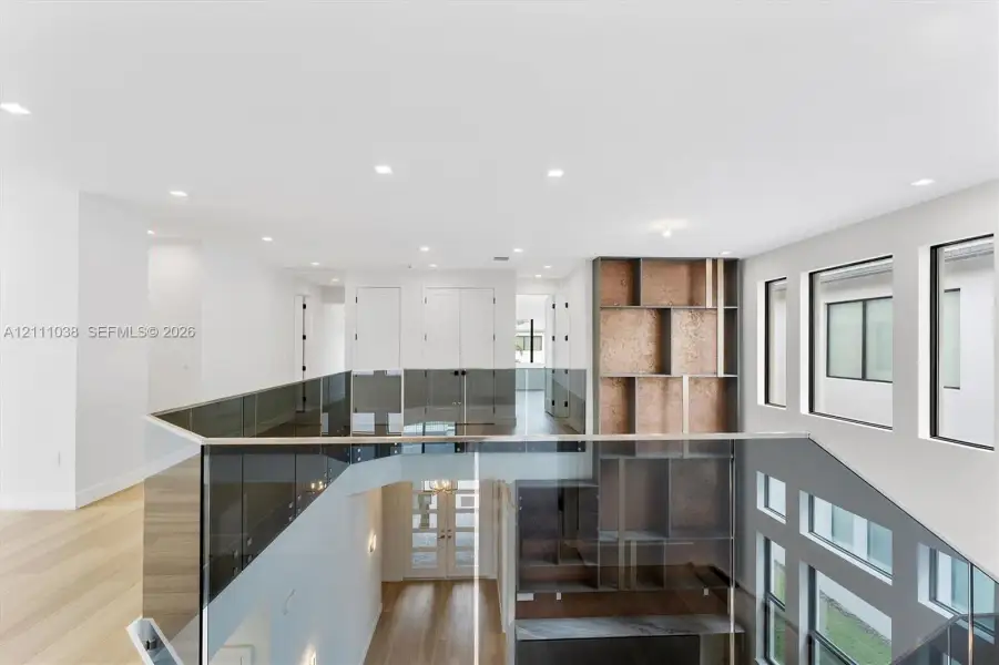 Open landing featuring wood-finish flooring, recessed lighting, floor-to-ceiling glass panel railing, and built-in vertical shelving with textured metallic paneling