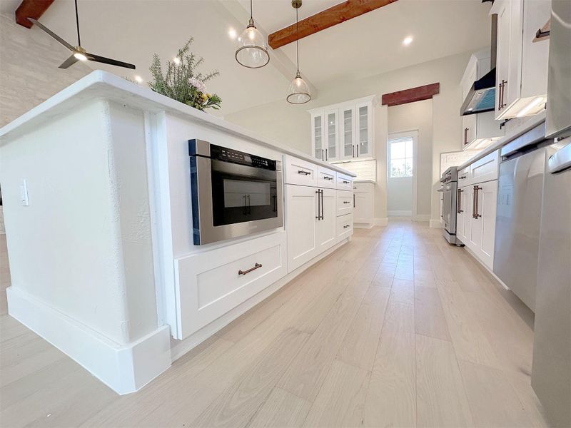 Kitchen with glass insert cabinets, white cabinets, counter microwave , all appliances with stainless steel finishes, light oak hardwood flooring, and pendant lighting