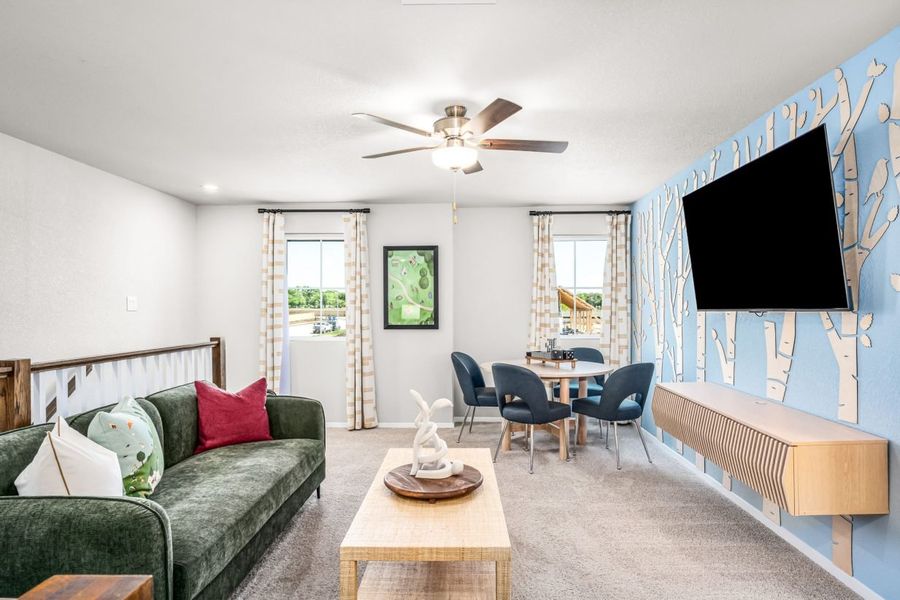 Image of a recreation room with a light blue wall with tree decoration, tan carpet, an accented wall with greenery, brown ceiling fan, with a table with black chairs, green couch with colorful pillows.