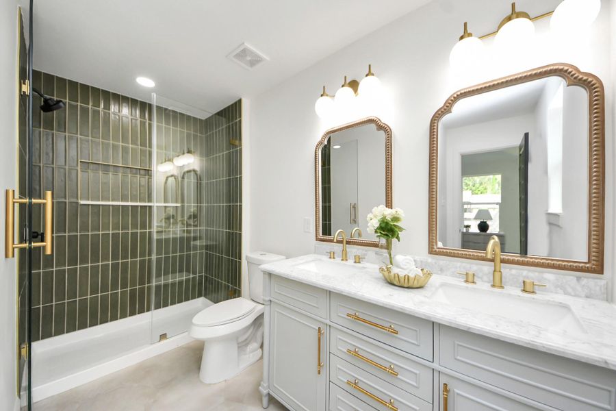 This bathroom features a sleek design with a modern glass-enclosed shower and stylish green tile. It includes a double vanity with elegant gold fixtures and large mirrors, providing a luxurious and functional space.