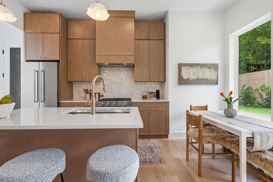 Contemporary kitchen featuring a large central island with a built-in sink and gold-tone faucet, light wood-finish cabinetry, stainless steel appliances, and light wood-finish flooring