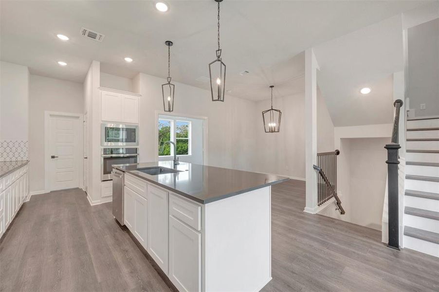 Open-concept kitchen featuring an oversized island with an undermount sink, built-in stainless steel appliances, white shaker cabinetry, and wood-finish flooring Open-concept kitchen featuring an oversized island with an undermount sink, built-in stainless steel appliances, white shaker cabinetry, and wood-finish flooring