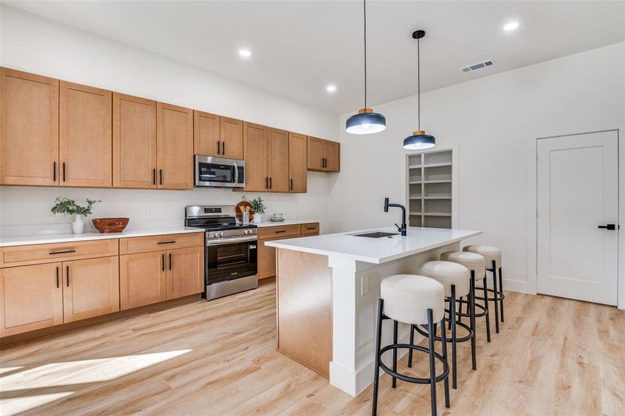 Kitchen featuring appliances with stainless steel finishes, decorative light fixtures, light wood-style floors, a breakfast bar area, and a center island with sink Kitchen featuring appliances with stainless steel finishes, decorative light fixtures, light wood-style floors, a breakfast bar area, and a center island with sink