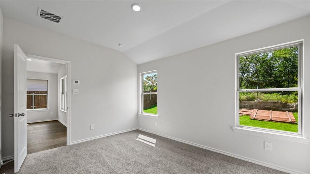 Carpeted room featuring two windows with outdoor views, light gray walls, and an open doorway leading to a room with wood-finish flooring