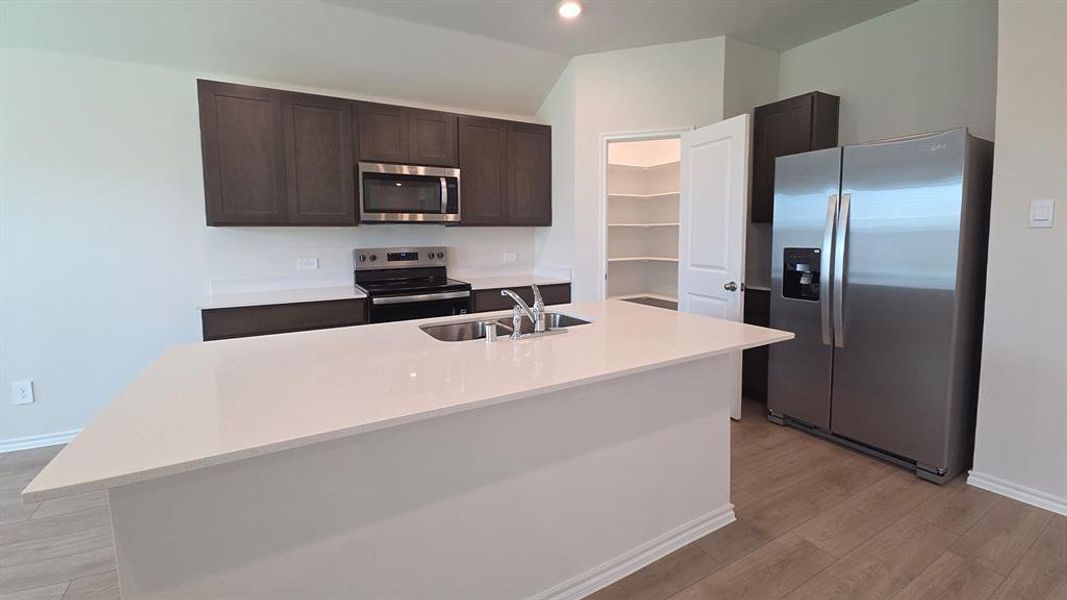 Modern kitchen featuring a large central island with an undermount sink, stainless steel appliances, dark wood-finish cabinetry, and light-toned countertops Modern kitchen featuring a large central island with an undermount sink, stainless steel appliances, dark wood-finish cabinetry, and light-toned countertops