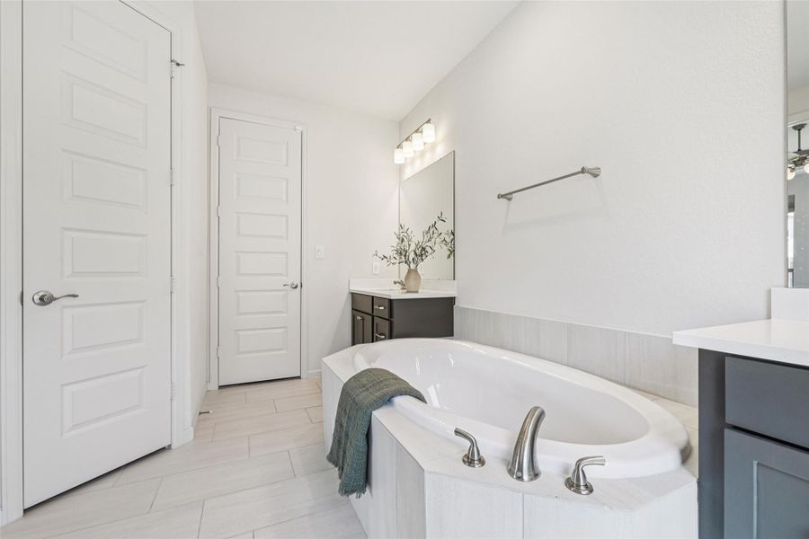 Bathroom featuring a spacious built-in soaking tub, a double vanity with dark cabinetry and light countertops, and light-colored tile flooring