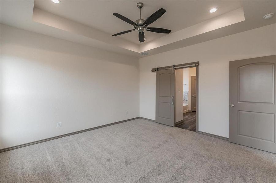 Unfurnished bedroom featuring a barn door, a raised ceiling, connected bathroom, carpet, and a ceiling fan