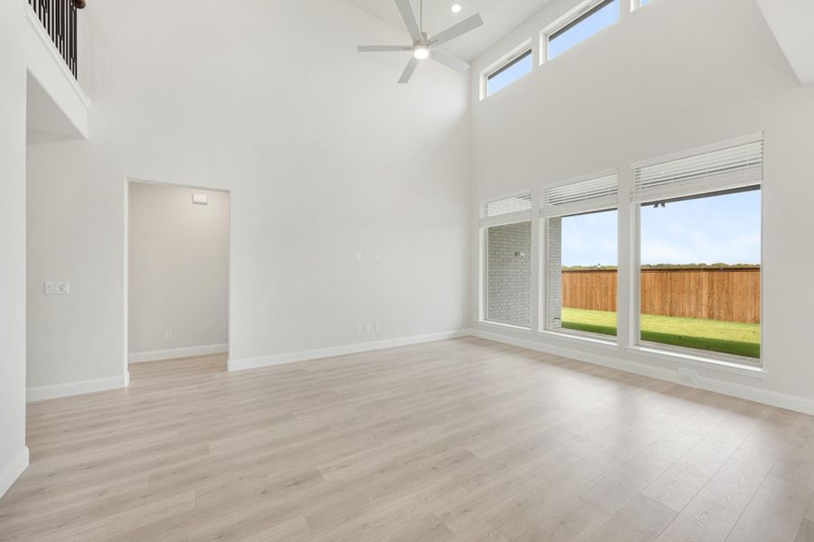Open living room with high vaulted ceiling, ceiling fan, light wood floors, and large windows overlooking backyard