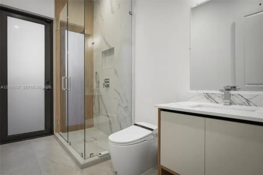 Full-height walk-in shower with glass enclosure, integrated tile niche, stone-surface vanity with undermount sink, linear wood-slat accent wall, and frosted glass door