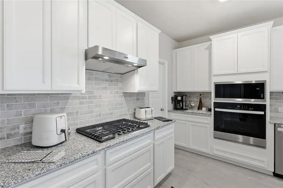Kitchen featuring white shaker-style cabinetry, stainless steel gas cooktop with coordinating vent hood, built-in microwave and oven combination, and granite countertops with a subway tile backsplash