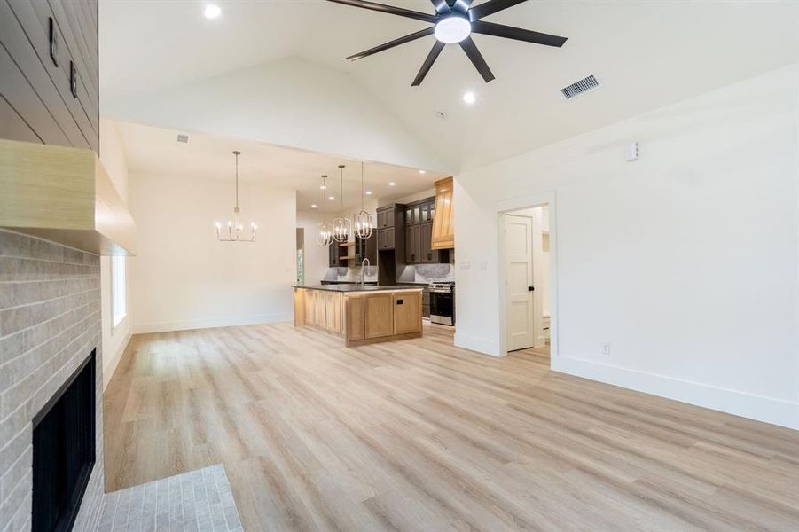 Unfurnished living room with a brick fireplace, ceiling fan with notable chandelier, sink, high vaulted ceiling, and light hardwood / wood-style flooring