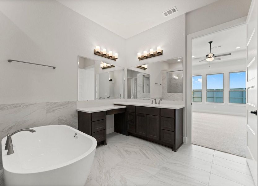 Awesome primary bathroom with a freestanding soaking tub,dual vanities w/large mirrors & elegant lighting.The space features sleek, dark cabinetry and light tiles, with an adjacent view into a bright,carpeted room with large windows and a ceiling fan.