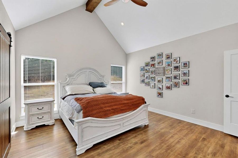 Bedroom featuring a barn door, high vaulted ceiling, beam ceiling, a ceiling fan, and light wood-style floors