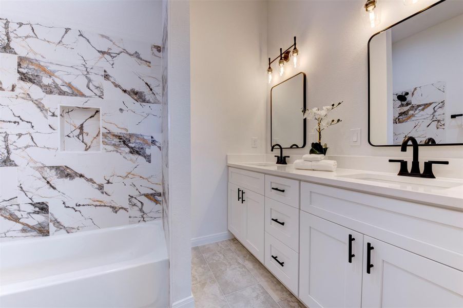 Secondary bathroom with double vanity, white shaker cabinets, quartz countertop, matte black fixtures, and floor-to-ceiling marble-look tile surround with built-in niche on tub/shower combo.