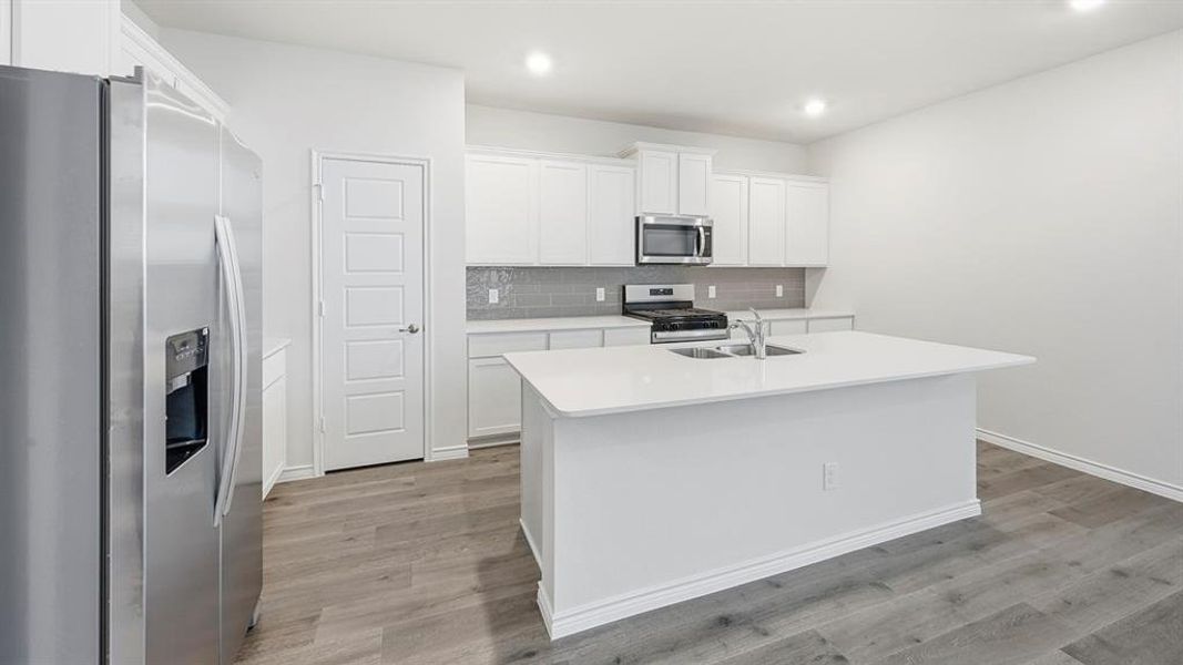 Modern kitchen featuring a central island with an integrated sink, stainless steel appliances, white cabinetry, gray subway tile backsplash, and wood-finish flooring