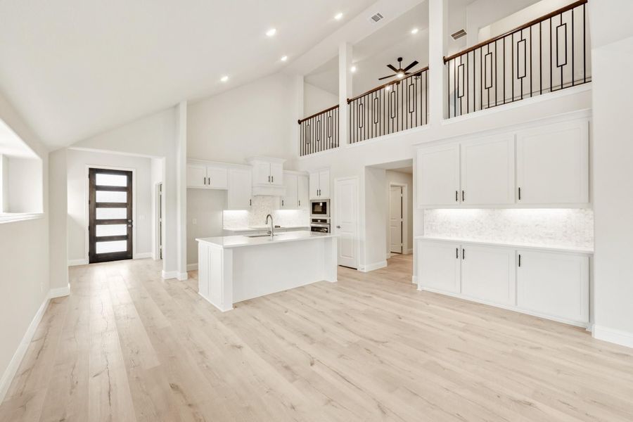 Open-concept kitchen with white cabinets, center island, and light wood floors under a two-story ceiling with iron railing balcony above.