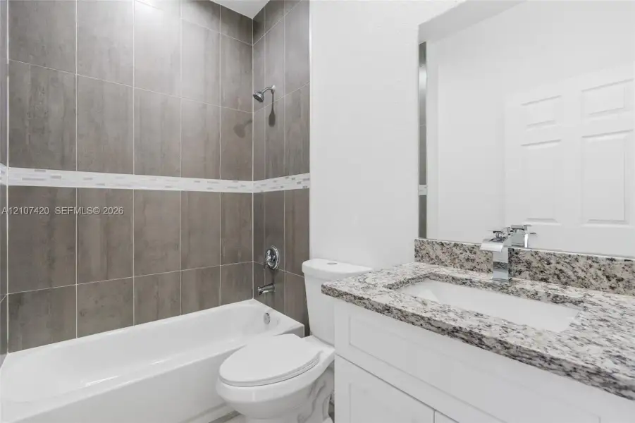 Bathroom featuring a vanity with a stone countertop, an integrated rectangular sink with a modern chrome faucet, a white toilet, and a bathtub with a shower head and full-height wall tile surround with decorative accent tiling Bathroom featuring a vanity with a stone countertop, an integrated rectangular sink with a modern chrome faucet, a white toilet, and a bathtub with a shower head and full-height wall tile surround with decorative accent tiling